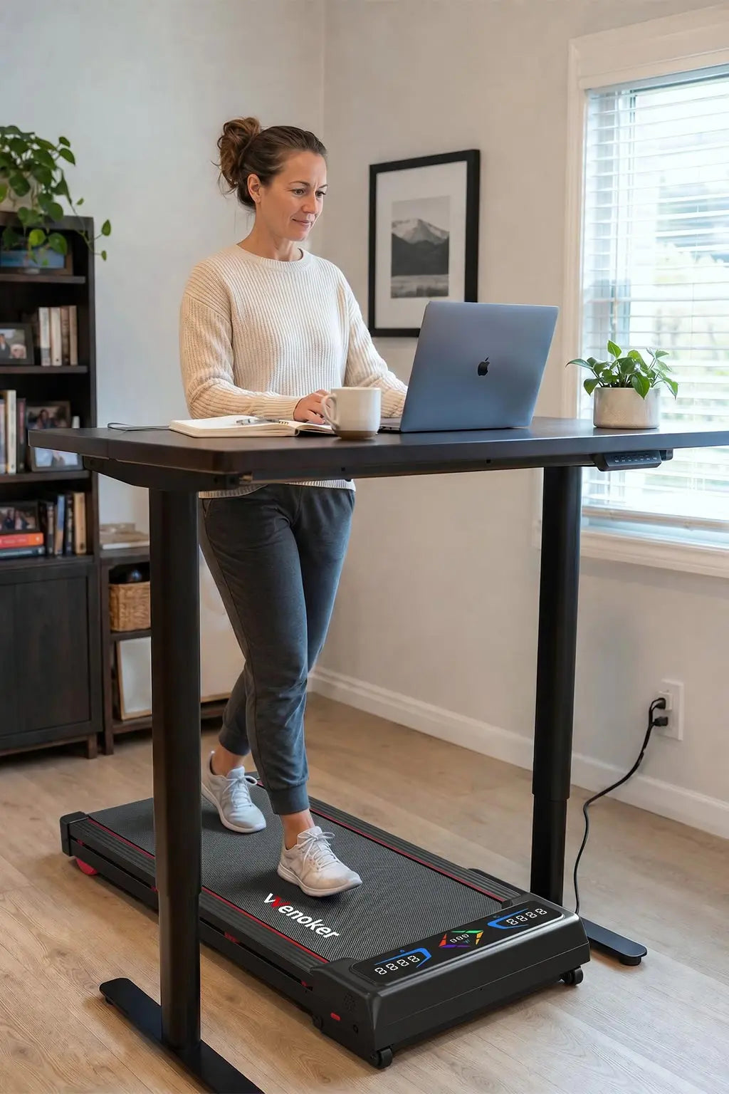 A woman working on a laptop at a standing desk while walking on a Wenoker under-desk walking pad treadmill in a bright home office.